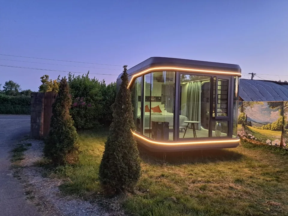 A pod at dusk with a glowing light line around its glazing, among conifers