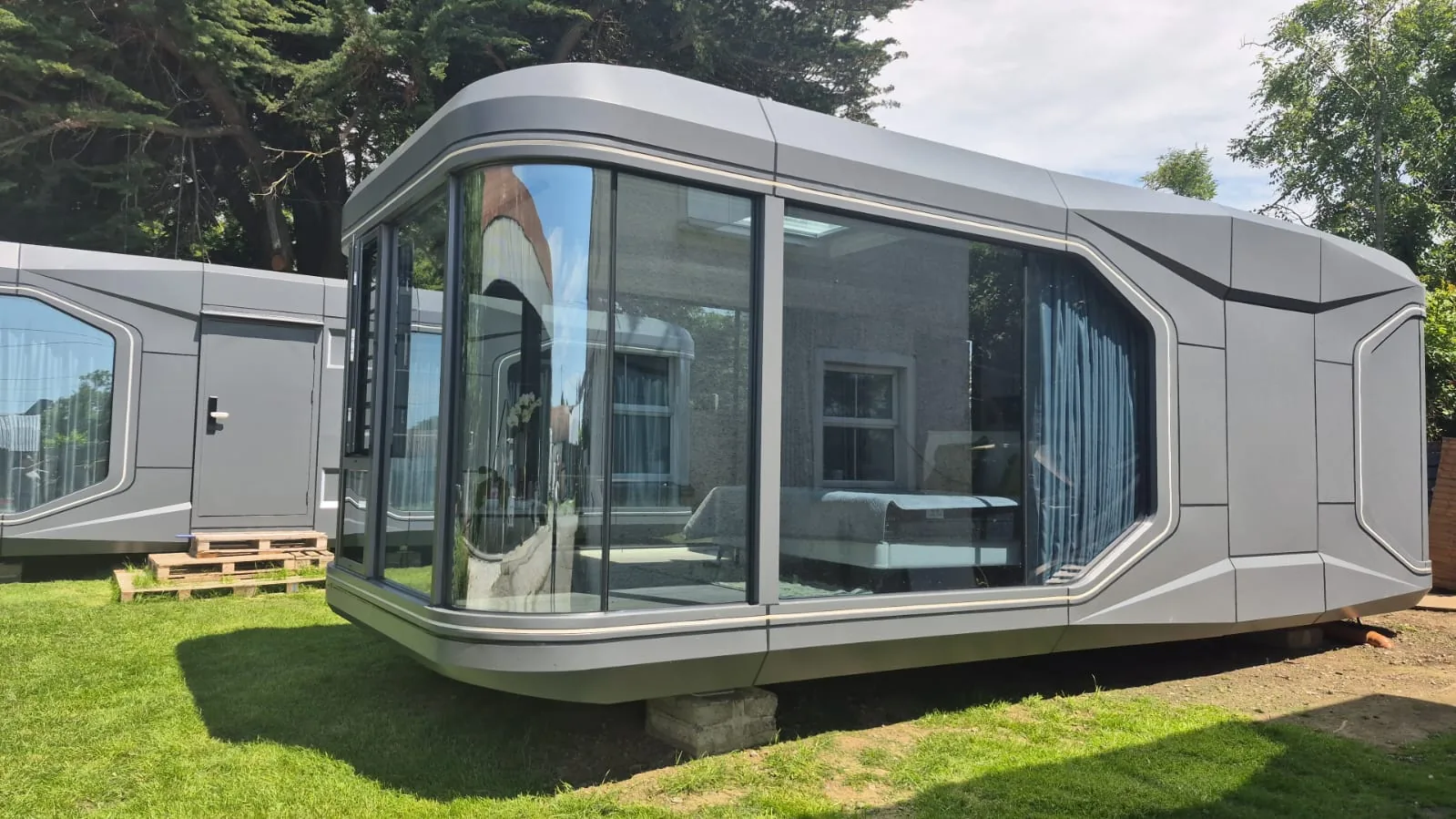 A grey aluminium pod with full-height curved glazing, set on a lawn beside a hedge