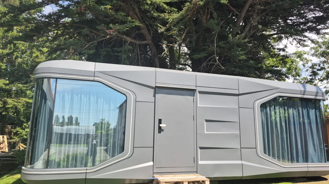 A long grey pod with a side door and curtained glazing at both ends, on a lawn
